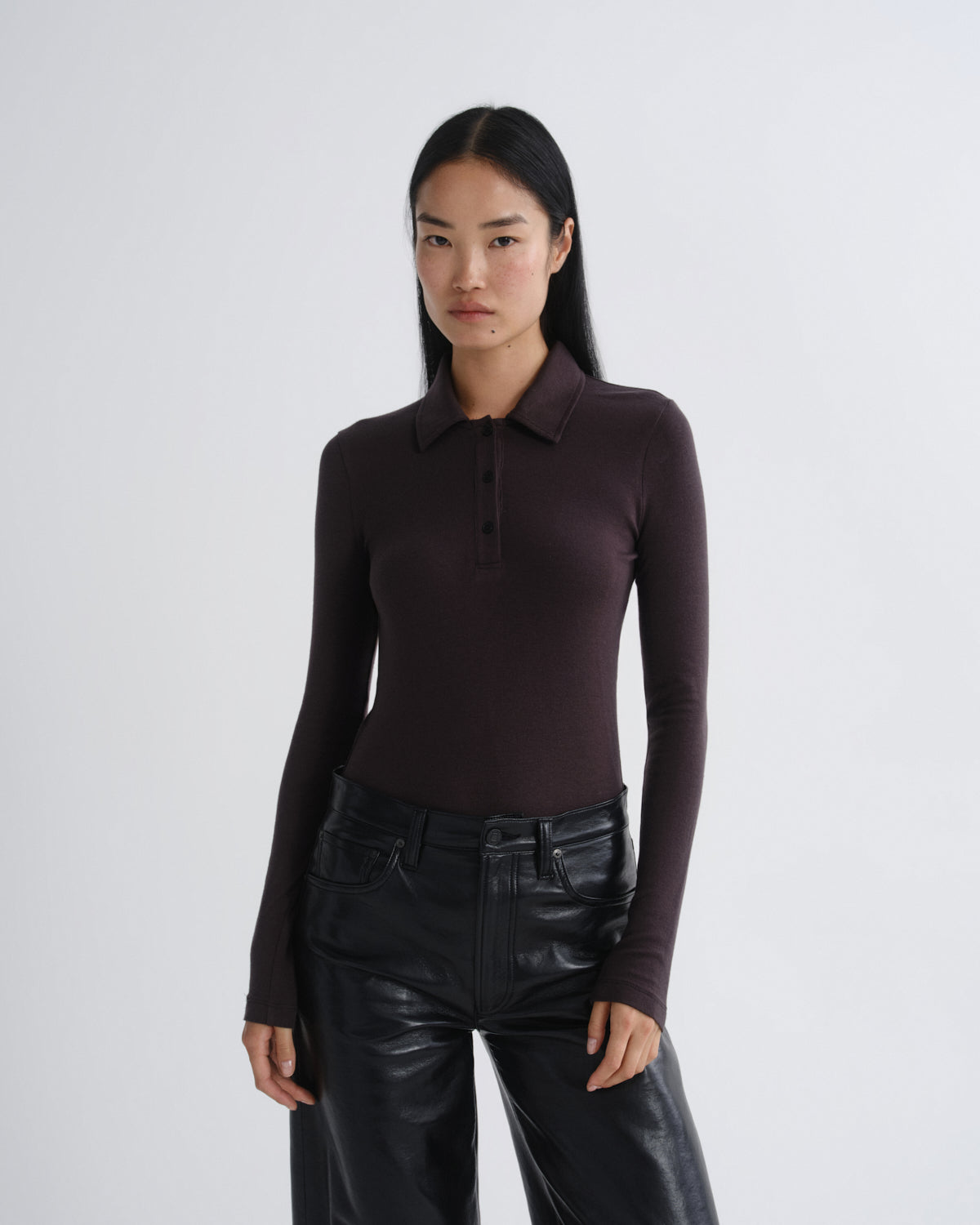A woman modeling a dark plum long-sleeve polo shirt paired with black leather pants, shot against a plain light gray studio background.