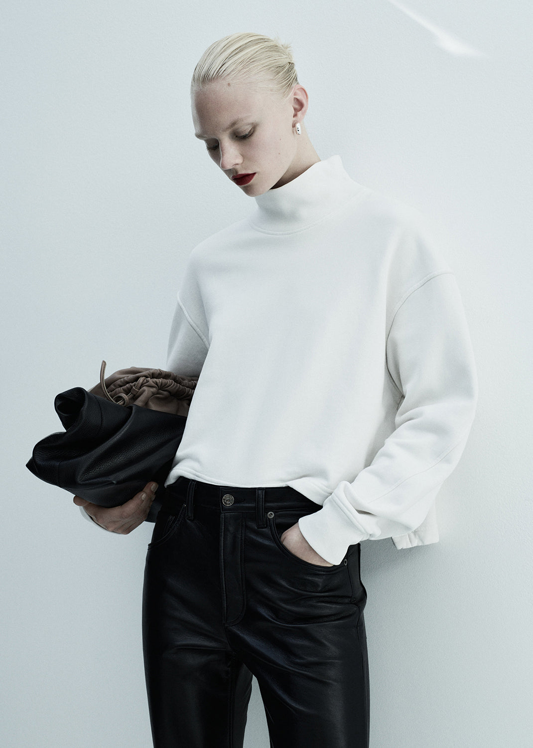 Person wearing a white sweater and black pants holding a black bag against a light background