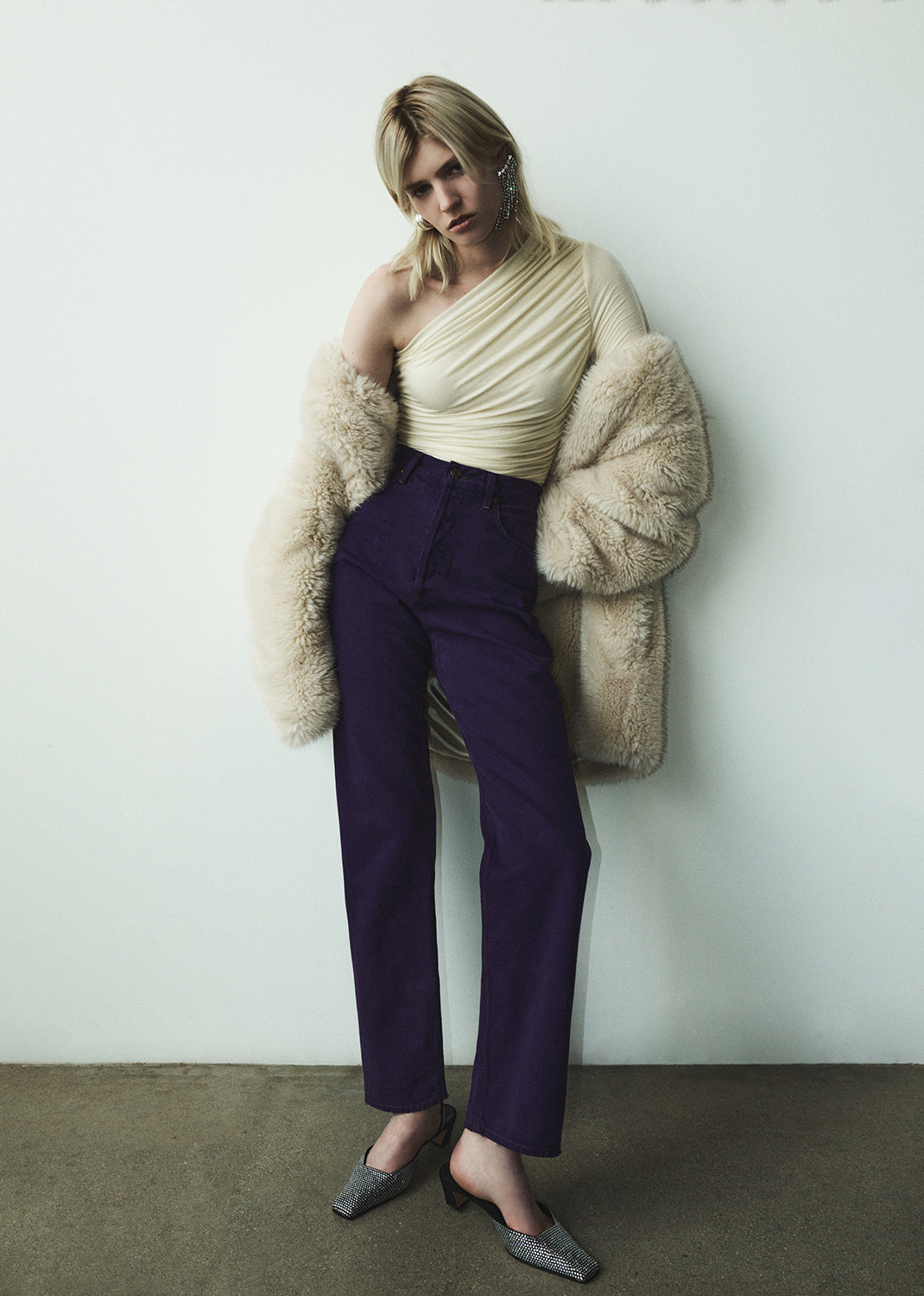 Woman wearing a cream top, purple pants, and a fur coat against a white wall.