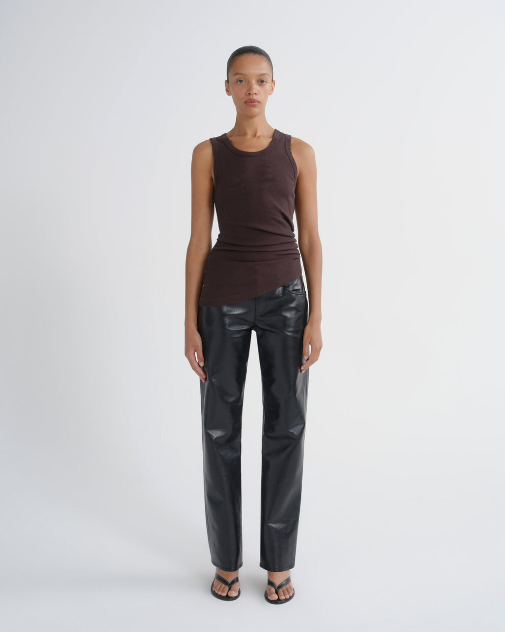 A model stands against a light gray studio background wearing a sleeveless dark brown top, shiny black faux-leather pants, and black sandals.
