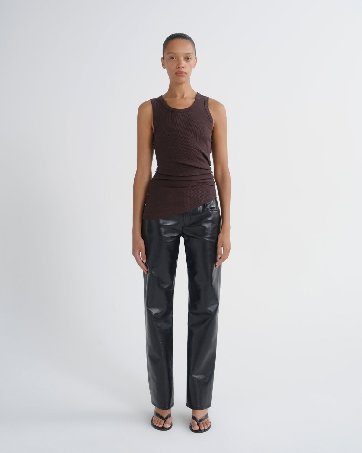 A model stands against a light gray studio background wearing a sleeveless dark brown top, shiny black faux-leather pants, and black sandals.