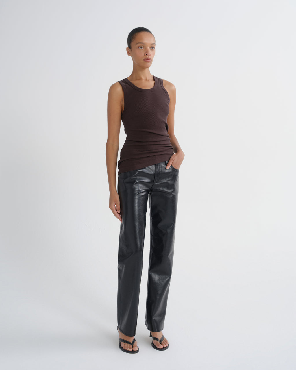 A model stands against a light gray studio background wearing a dark brown ribbed sleeveless top, black faux-leather straight-leg pants, and black sandals.