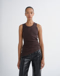 A model wearing a dark brown ribbed sleeveless tank top and black leather pants, standing against a light gray studio background.
