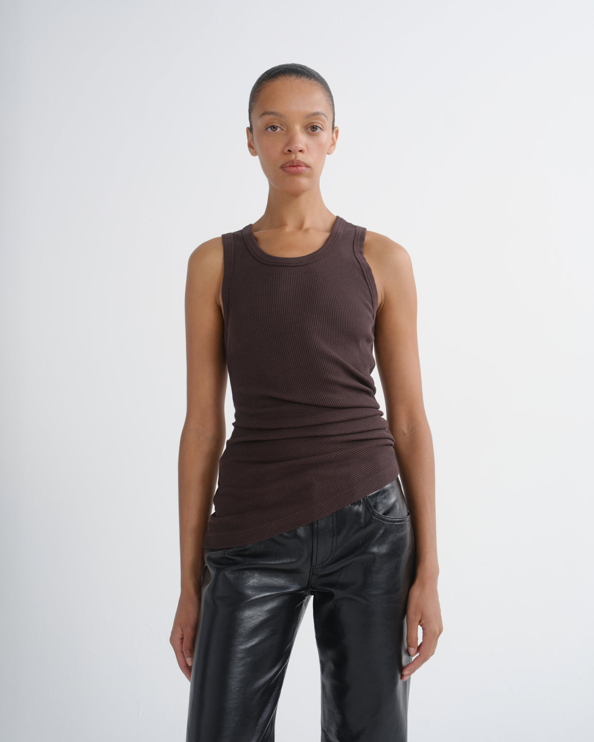 A model wearing a dark brown ribbed sleeveless tank top and black leather pants, standing against a light gray studio background.