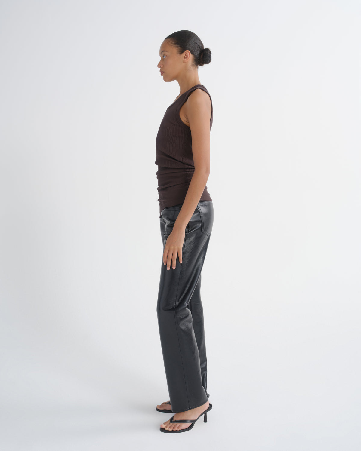 A woman stands in profile wearing a dark brown sleeveless top, shiny dark gray wide-leg pants, and black heeled sandals against a plain light gray studio background.