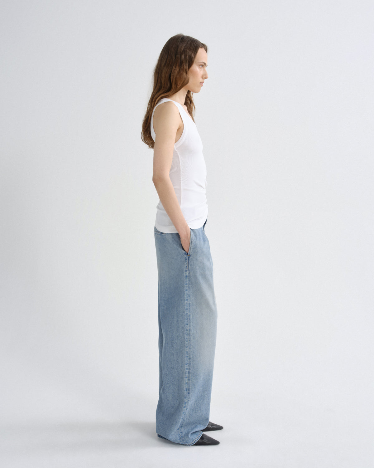 A woman stands in profile wearing a white ribbed tank top and light-wash wide-leg denim jeans against a plain light gray studio background.