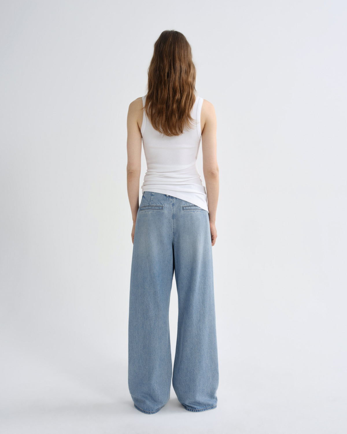 A woman viewed from behind wearing a white ribbed tank top and light-wash wide-leg denim jeans against a plain light gray studio background.