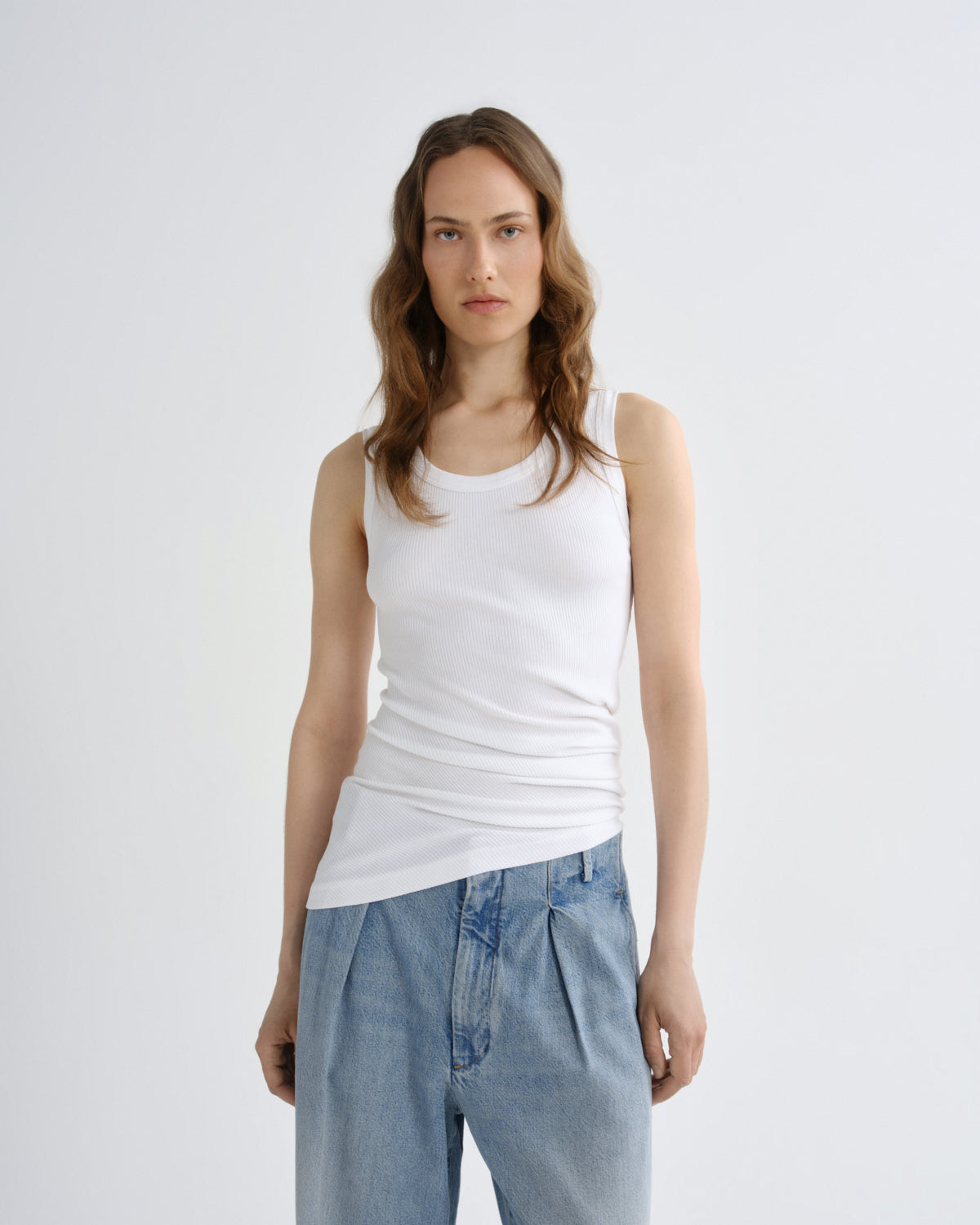 A woman modeling a white ribbed tank top paired with light-wash blue jeans, standing against a plain light gray studio background.