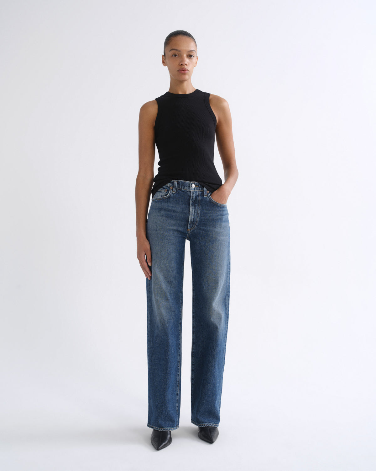 A model stands against a plain light gray studio background wearing a sleeveless black top and straight-leg blue jeans.