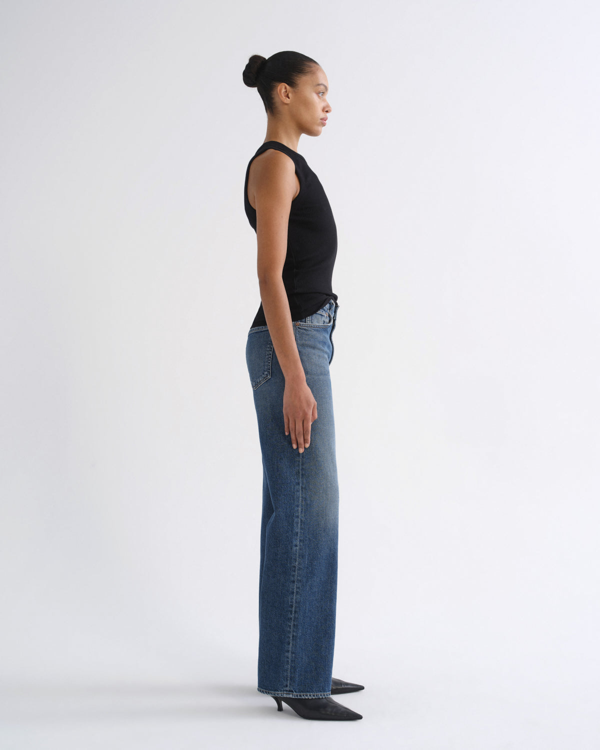 A model in profile wearing a black sleeveless top and wide-leg blue jeans, standing against a plain light gray studio background.