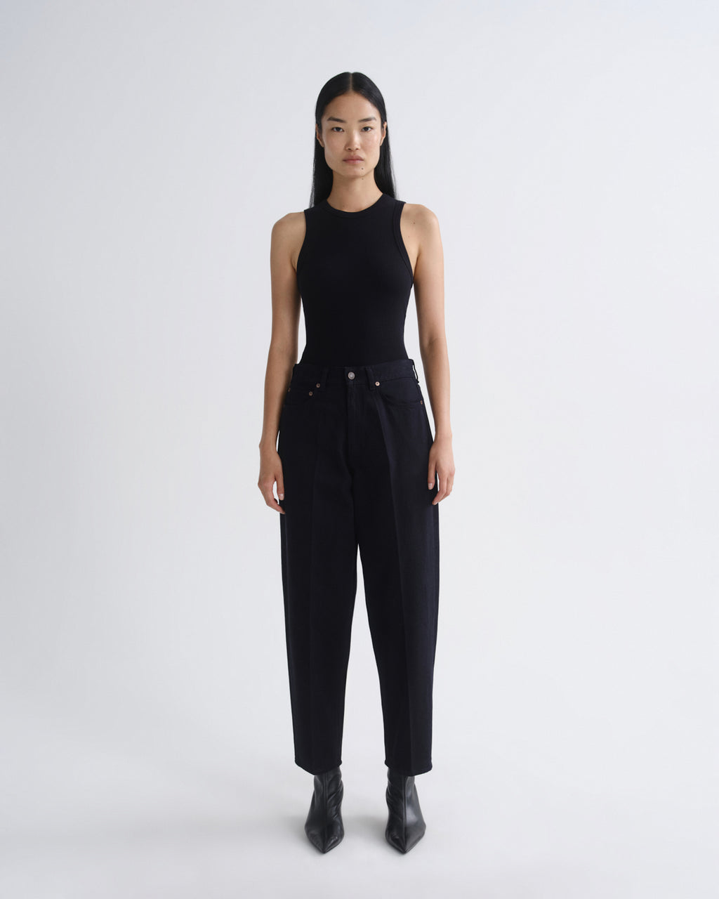 A model stands against a light gray studio background wearing a black sleeveless mock-neck top and black high-waisted wide-leg trousers, paired with black pointed ankle boots.