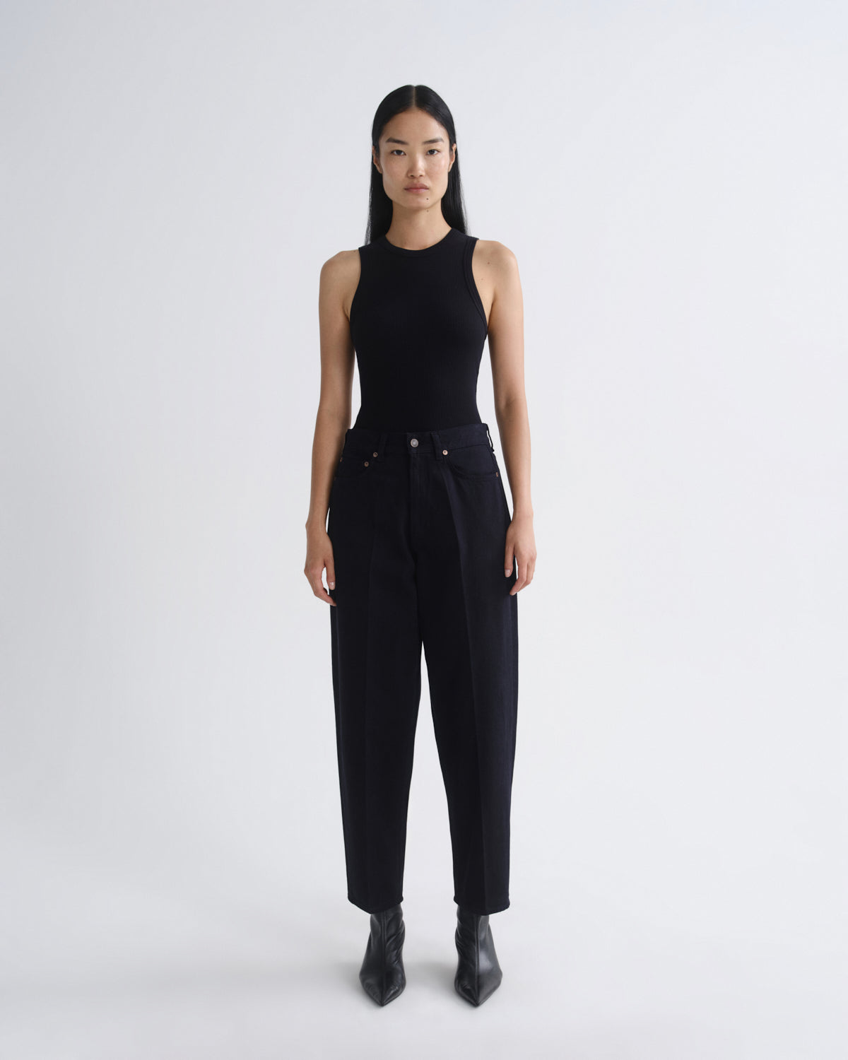 A model stands against a light gray studio background wearing a black sleeveless mock-neck top and black high-waisted wide-leg trousers, paired with black pointed ankle boots.