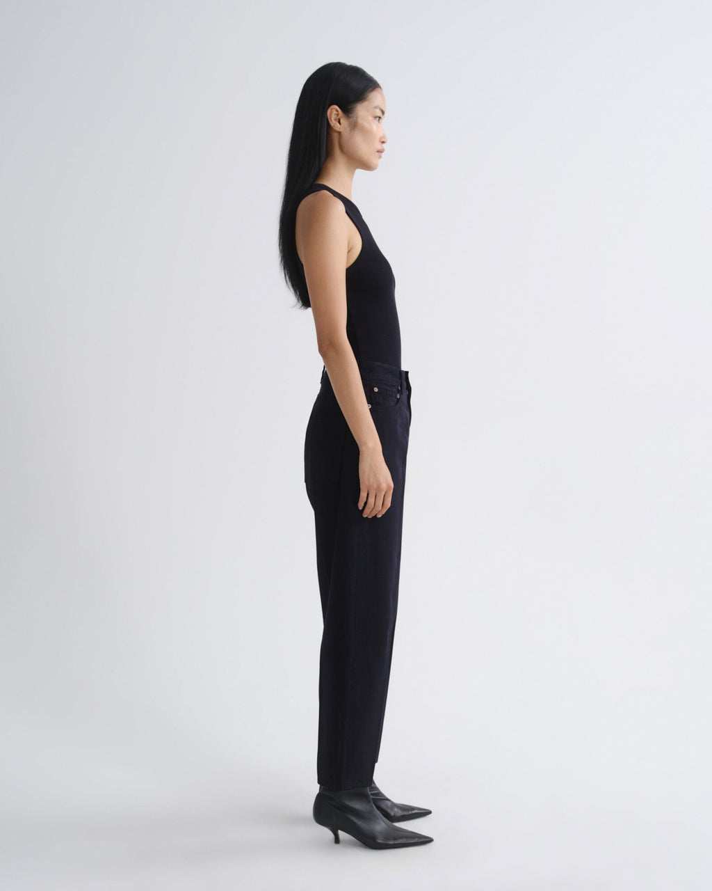 A model in a side profile view wearing a black sleeveless top and black wide-leg trousers, paired with black pointed-toe ankle boots, against a plain light gray studio background.