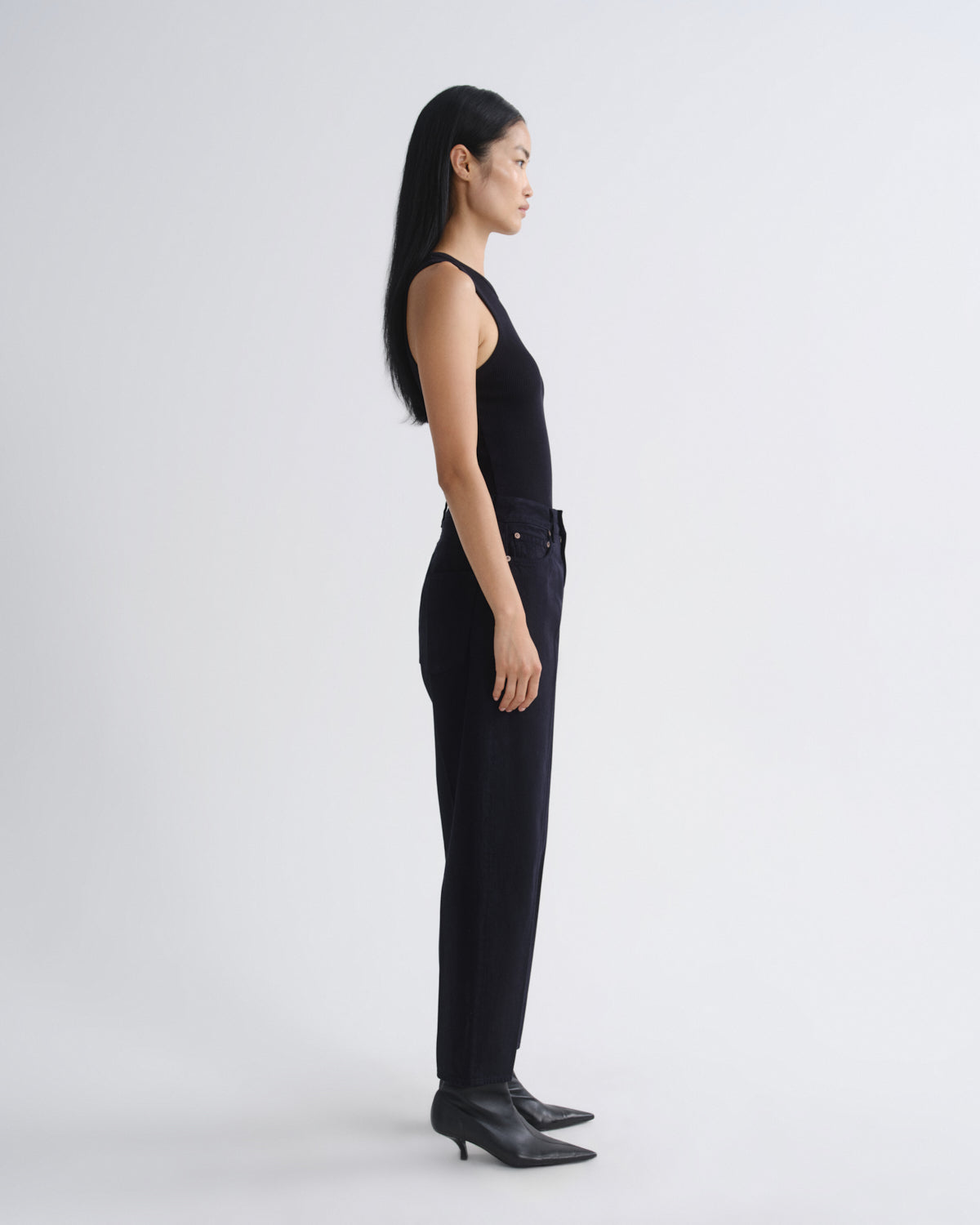 A model in a side profile view wearing a black sleeveless top and black wide-leg trousers, paired with black pointed-toe ankle boots, against a plain light gray studio background.
