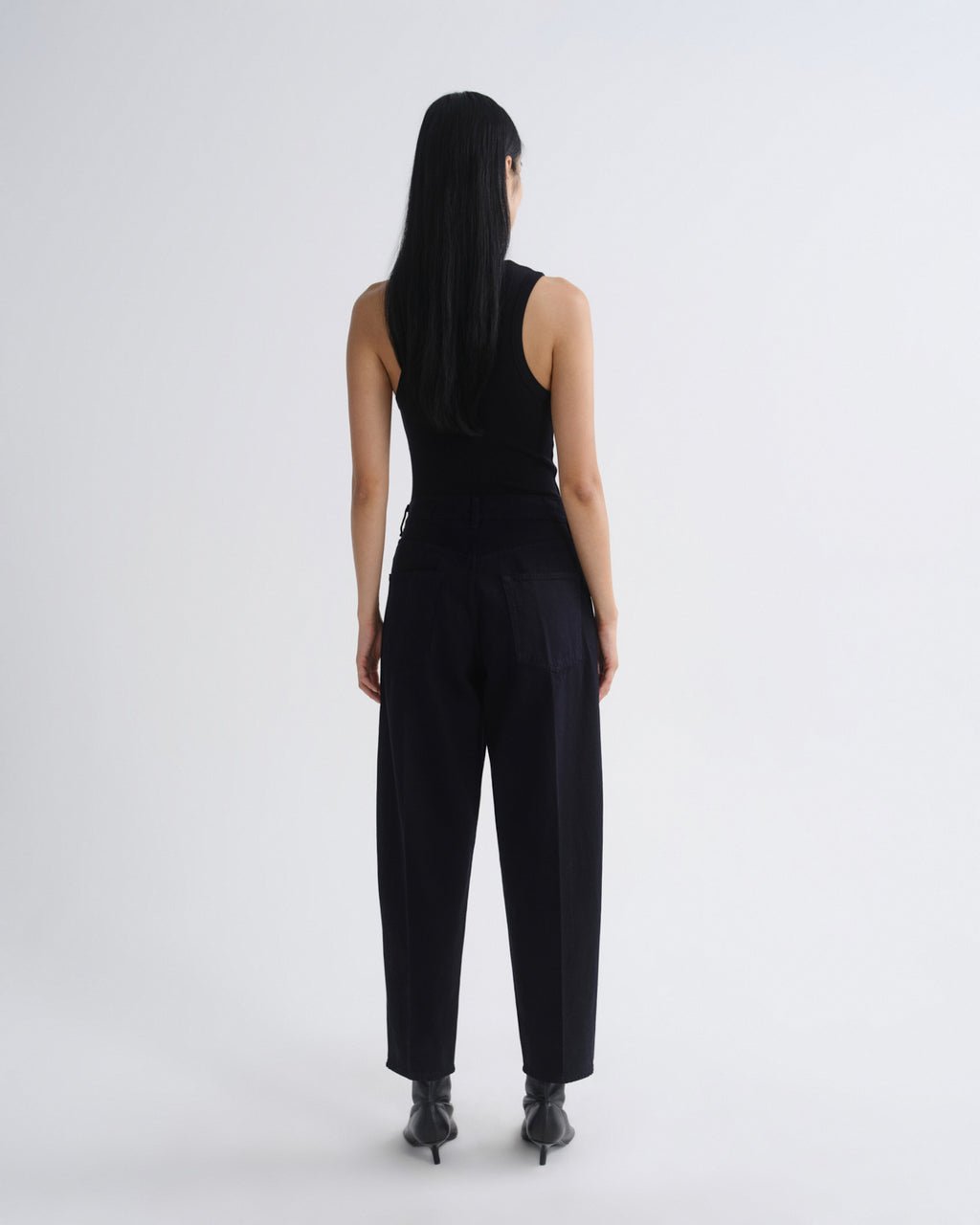 A model viewed from behind wearing a black sleeveless top and black wide-leg trousers, standing against a plain light gray studio background.