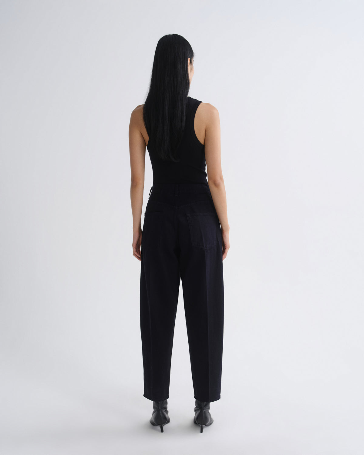 A model viewed from behind wearing a black sleeveless top and black wide-leg trousers, standing against a plain light gray studio background.