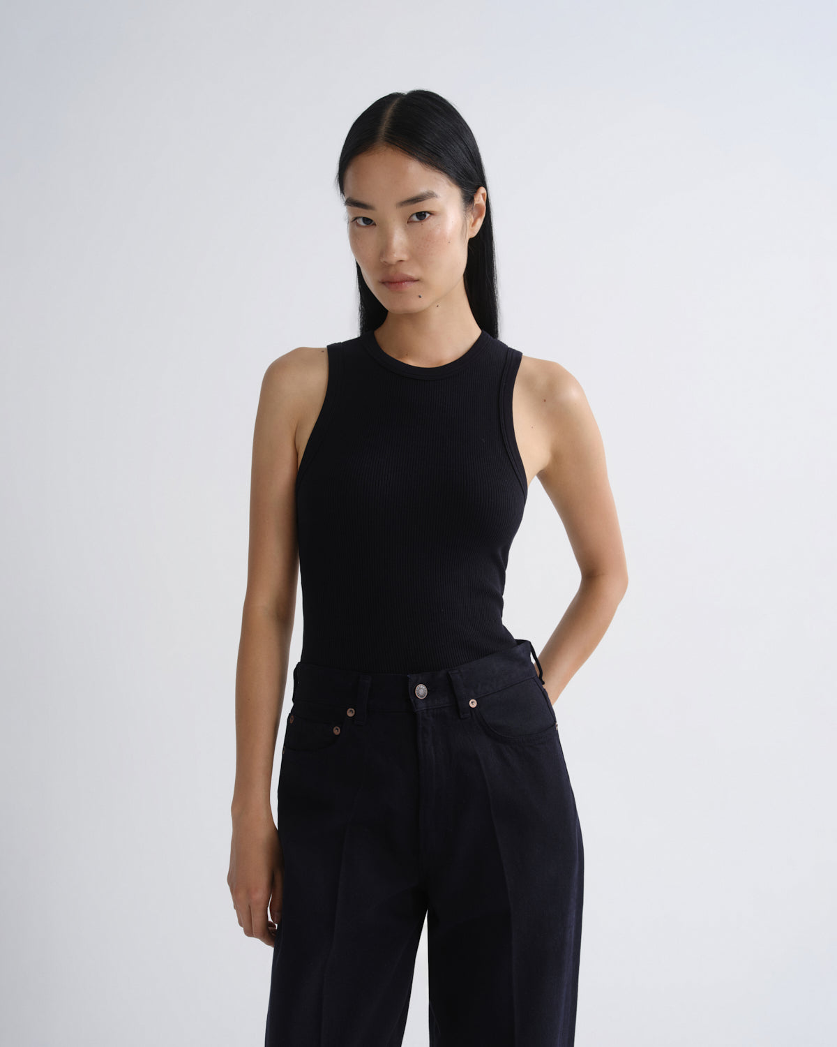 A model wearing a black sleeveless mock-neck top and black wide-leg jeans, standing against a light gray studio background.