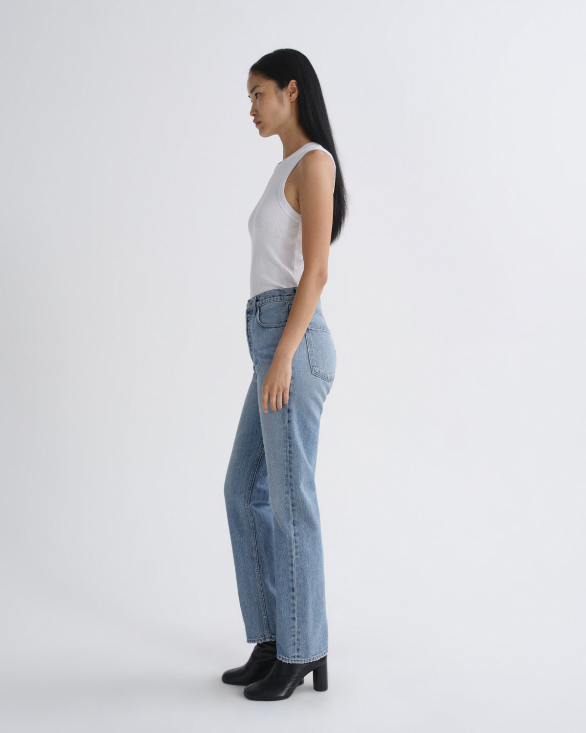A woman in a white sleeveless top and light blue wide-leg jeans stands in profile against a plain light gray studio background, wearing black ankle boots.