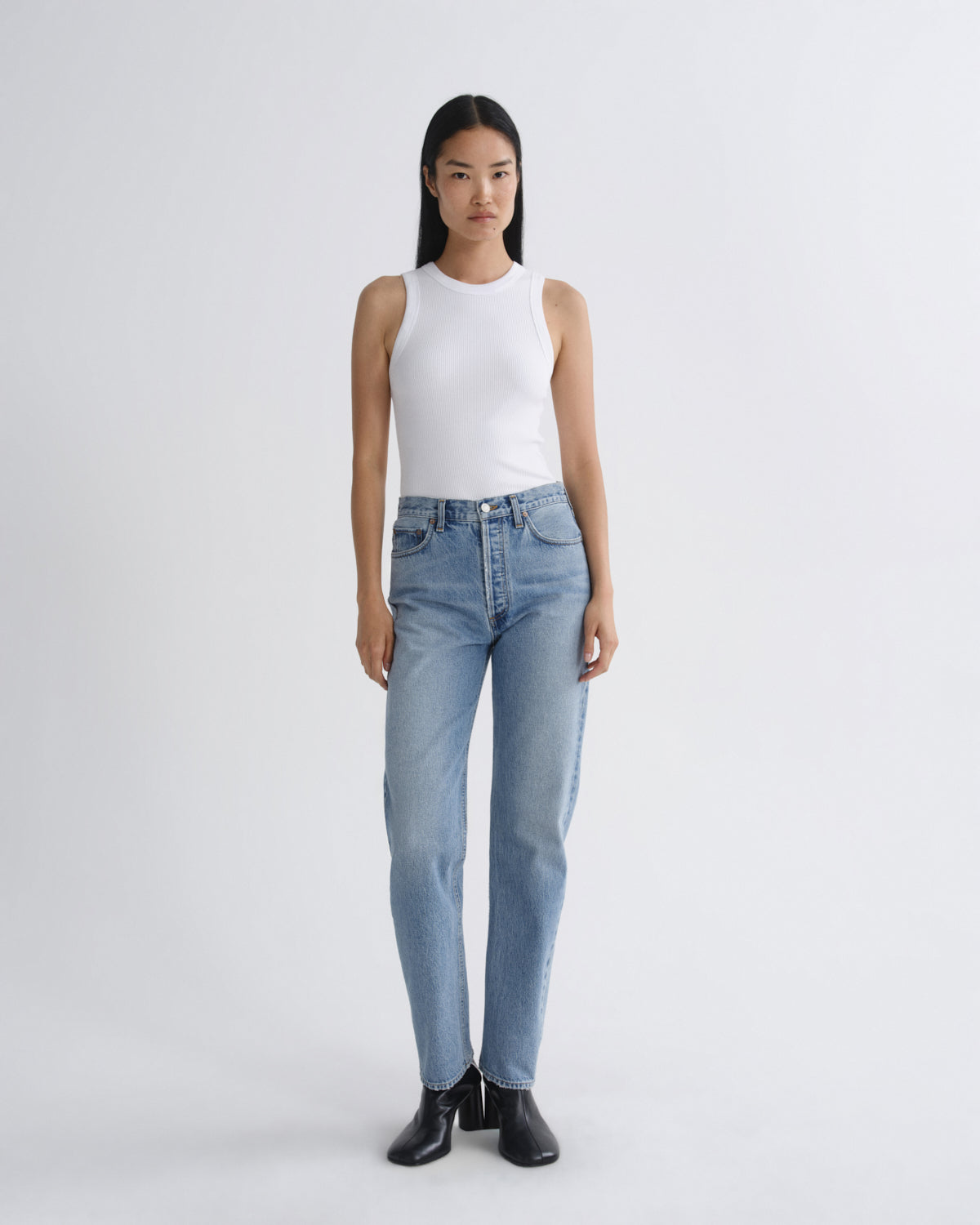 A model stands against a light gray studio background wearing a white sleeveless top, light-wash blue jeans, and black ankle boots.