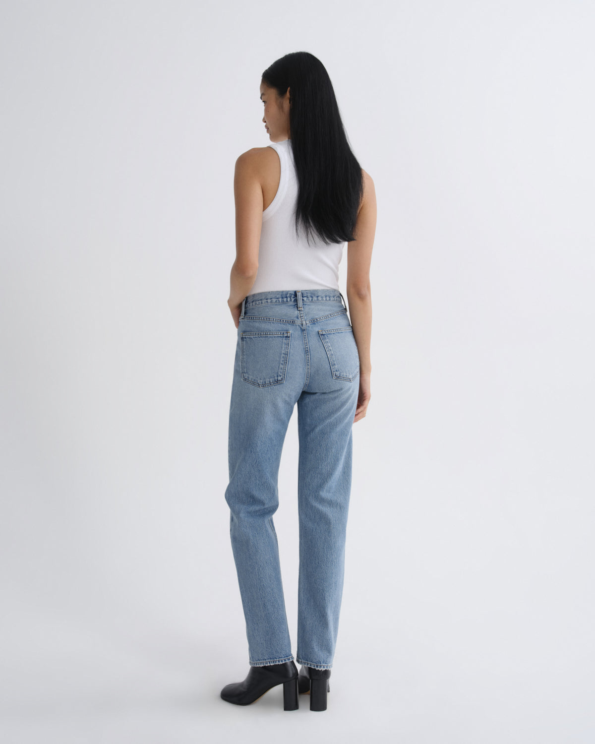 A woman with long black hair is shown from behind wearing light-wash blue jeans, a white sleeveless top, and black ankle boots against a plain light gray studio background.