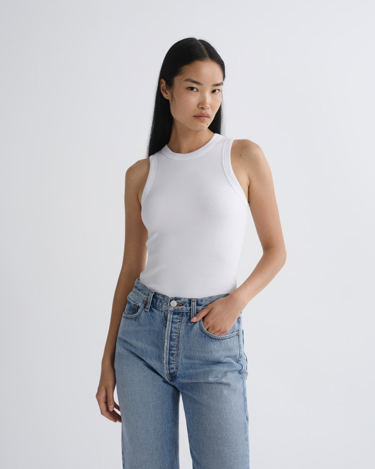 A woman modeling a white sleeveless ribbed tank top paired with light-wash blue jeans against a plain light gray studio background.