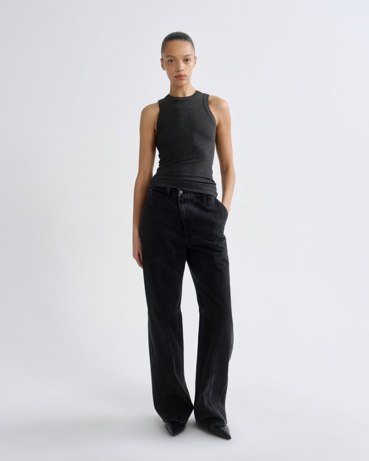 A model stands against a light gray studio background wearing a dark charcoal gray ribbed sleeveless mock-neck top and black wide-leg jeans.