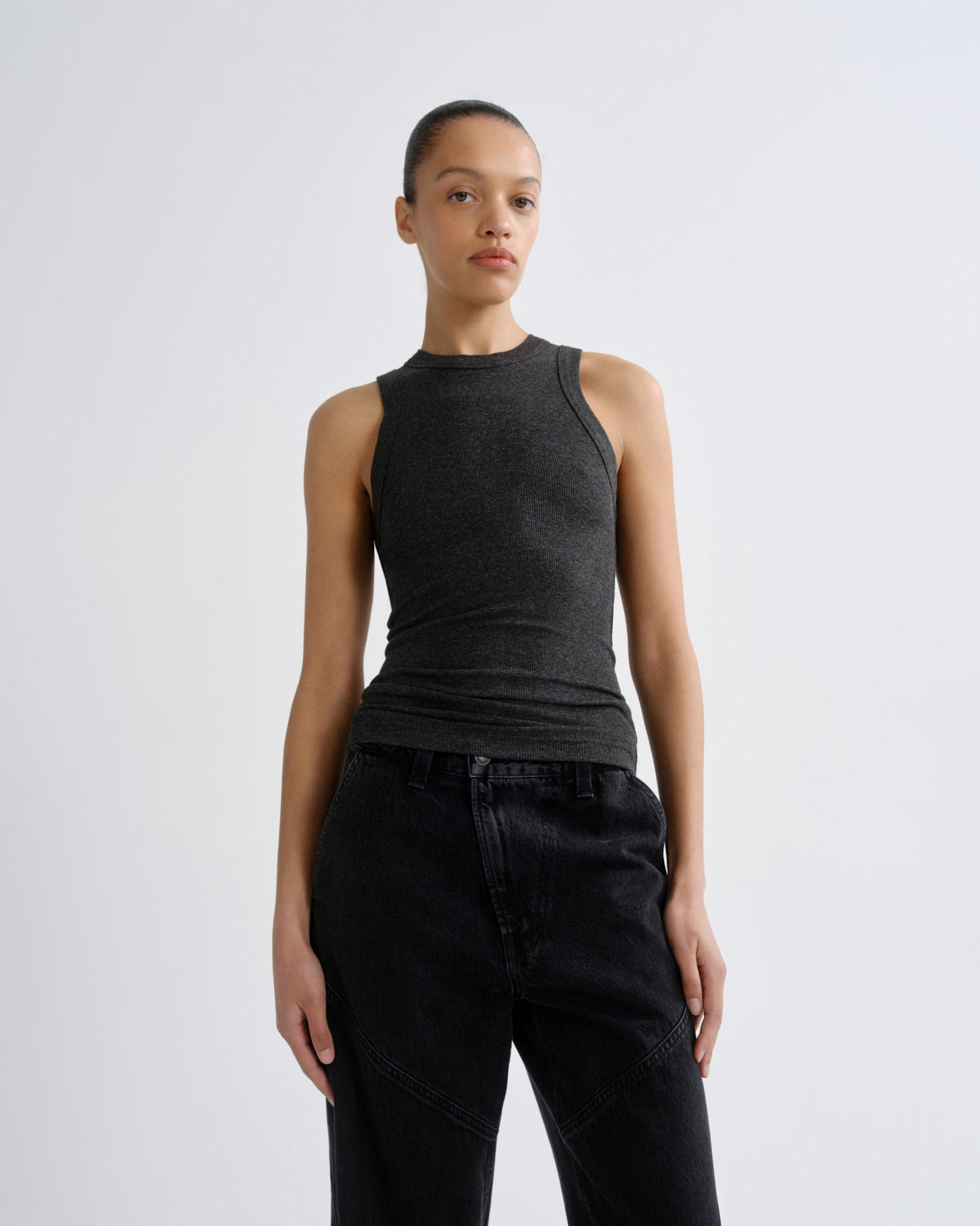 A model wearing a dark charcoal gray ribbed sleeveless mock neck top and black jeans, standing against a light gray studio background.