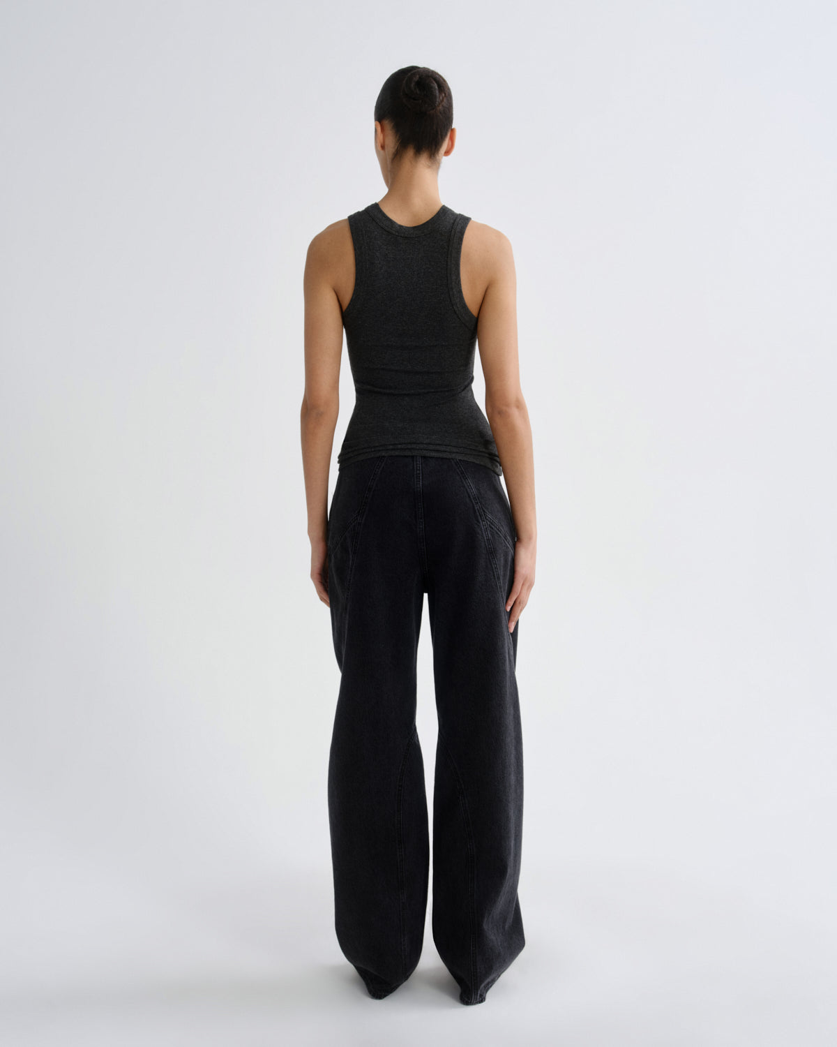 A model viewed from the back wearing a dark charcoal gray ribbed sleeveless top and black wide-leg trousers, standing against a light gray studio background.