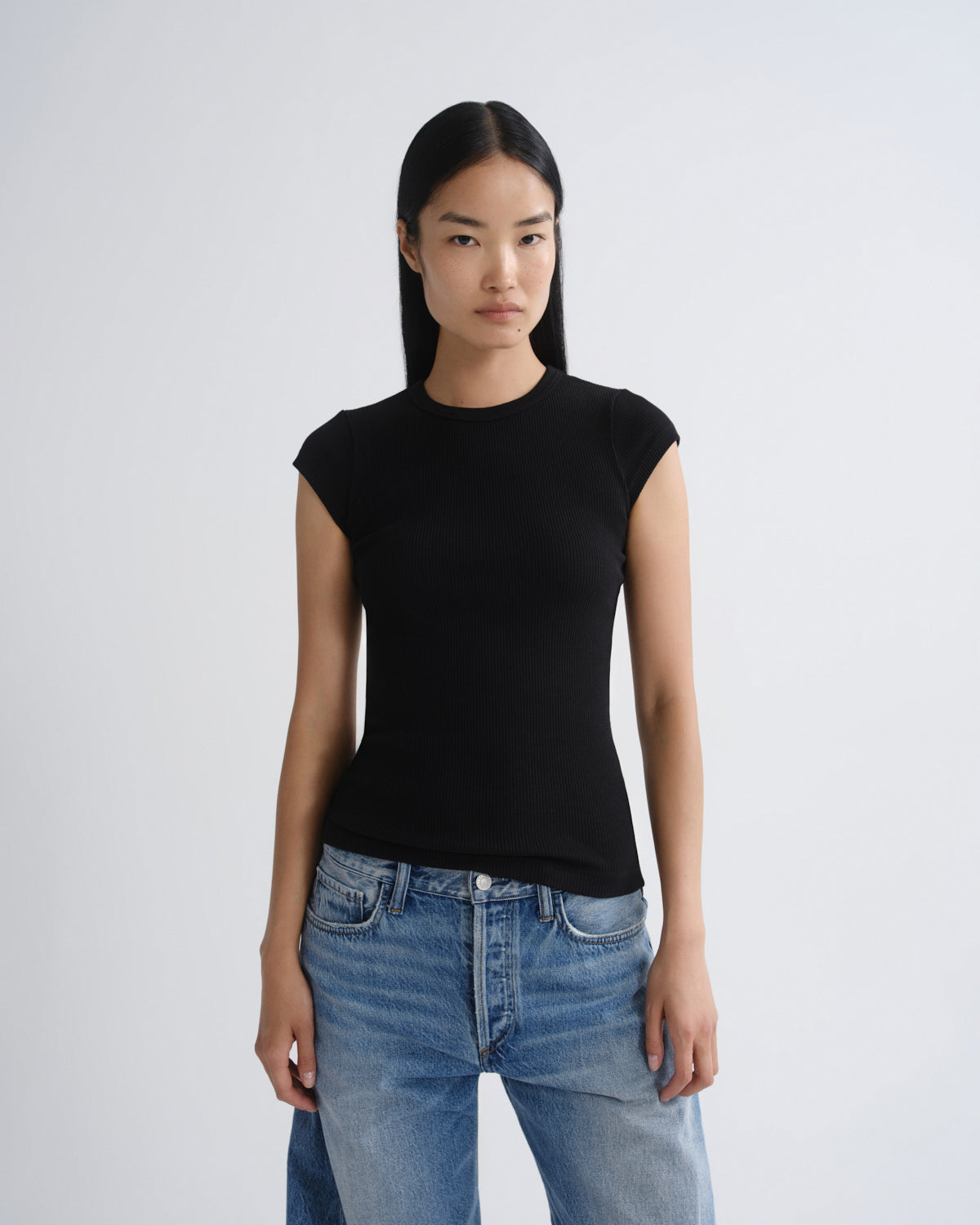 A model wearing a fitted black short-sleeve crewneck top and light wash blue jeans, standing against a light gray studio background.