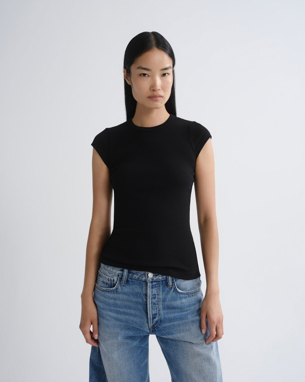 A model wearing a fitted black short-sleeve crewneck top and light wash blue jeans, standing against a light gray studio background.
