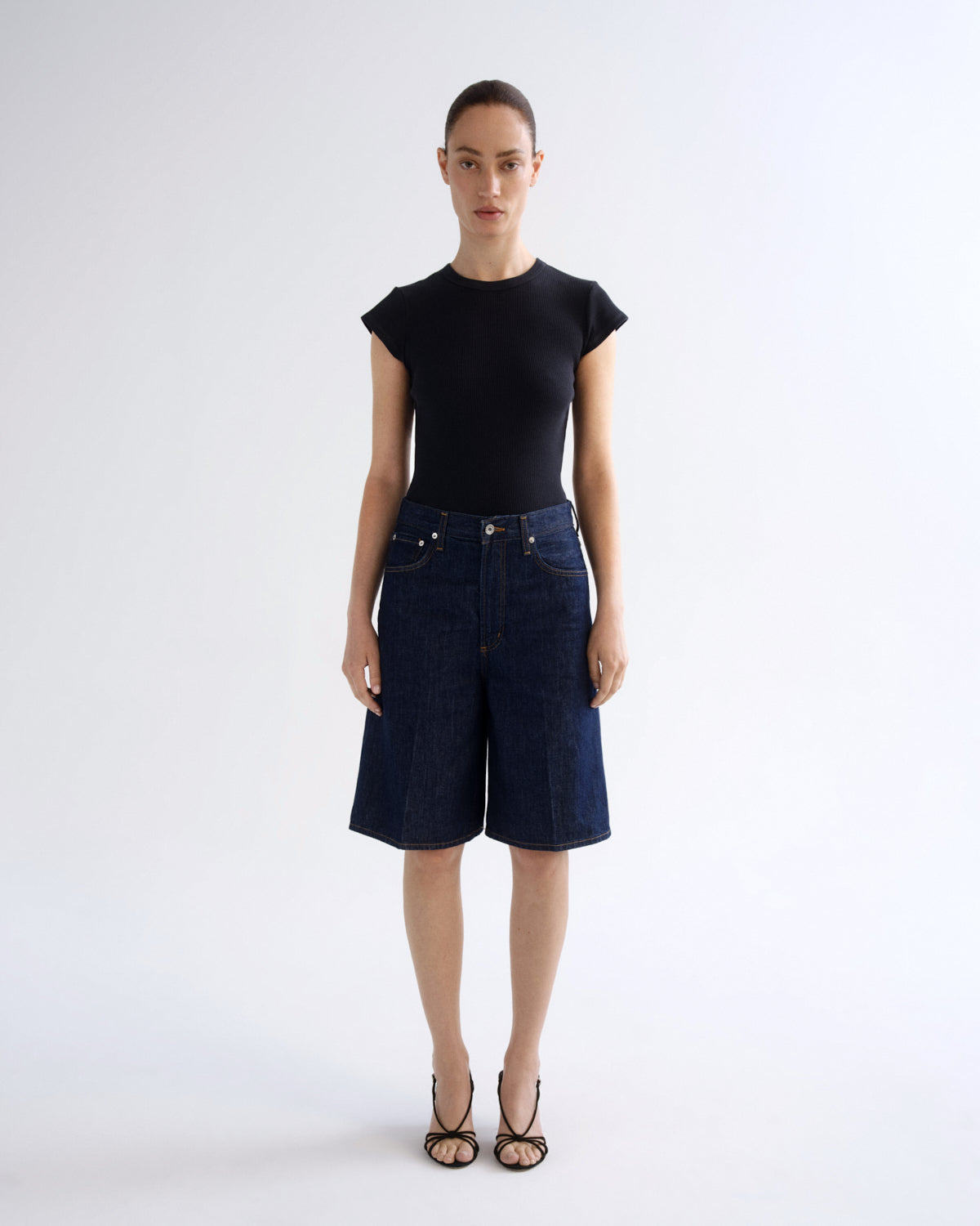 A model stands against a light gray studio background wearing a black short-sleeve top and dark blue denim shorts with black strappy heels.