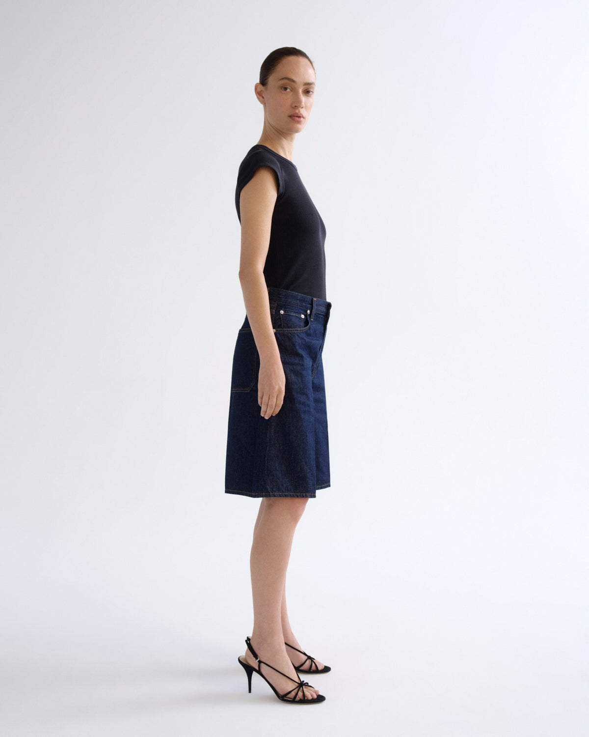 A model stands in profile wearing a dark navy blue denim skirt with front patch pockets, a black sleeveless top, and black strappy high-heeled sandals against a plain light gray studio background.