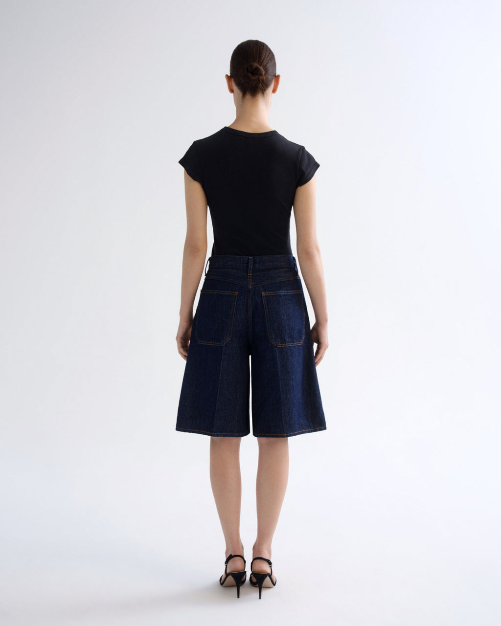 A model viewed from the back wearing a black short-sleeve top and dark blue denim culottes, paired with black high-heeled sandals, against a light gray studio background.
