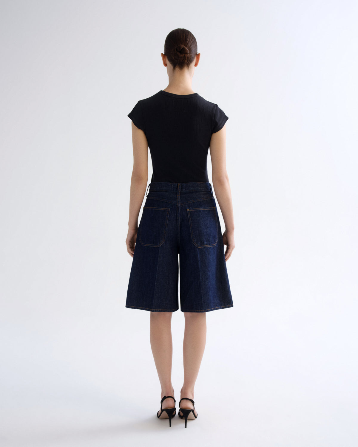 A model viewed from the back wearing a black short-sleeve top and dark blue denim culottes, paired with black high-heeled sandals, against a light gray studio background.