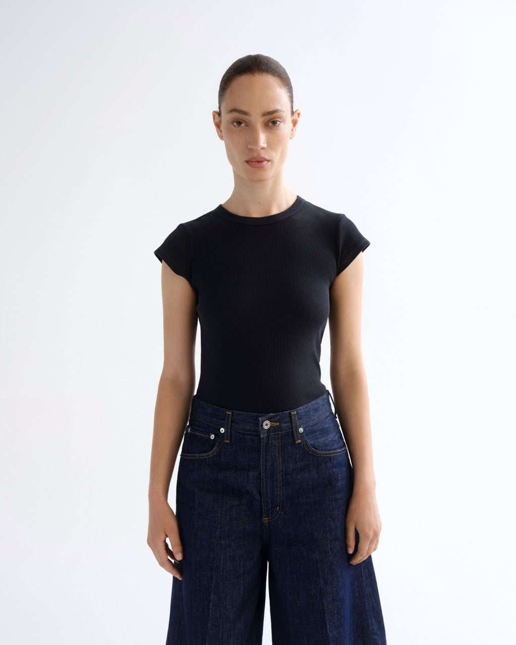 A model wearing a black short-sleeve crewneck top and dark blue wide-leg jeans, standing against a plain white studio background.