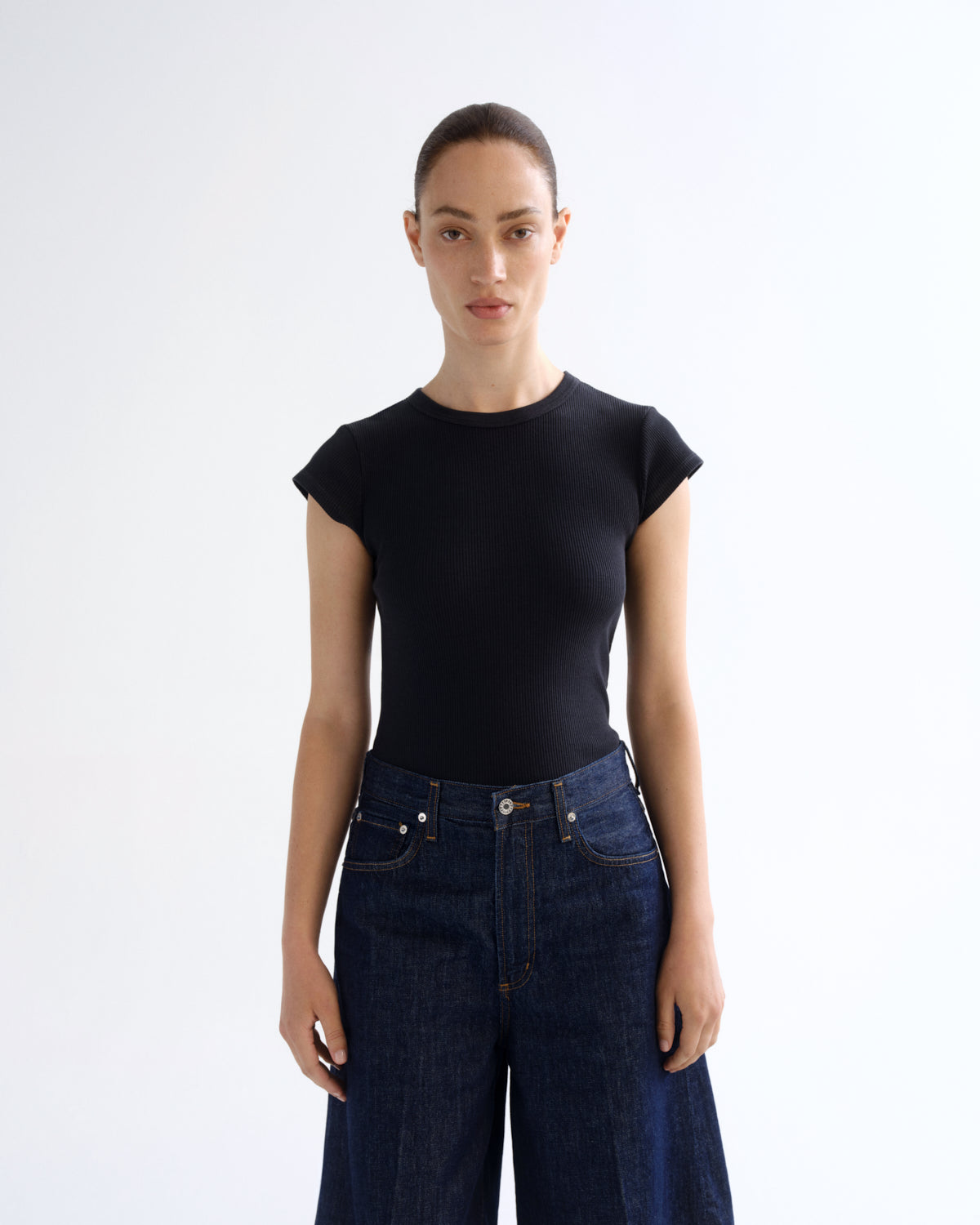 A model wearing a black short-sleeve crewneck top and dark blue wide-leg jeans, standing against a plain white studio background.