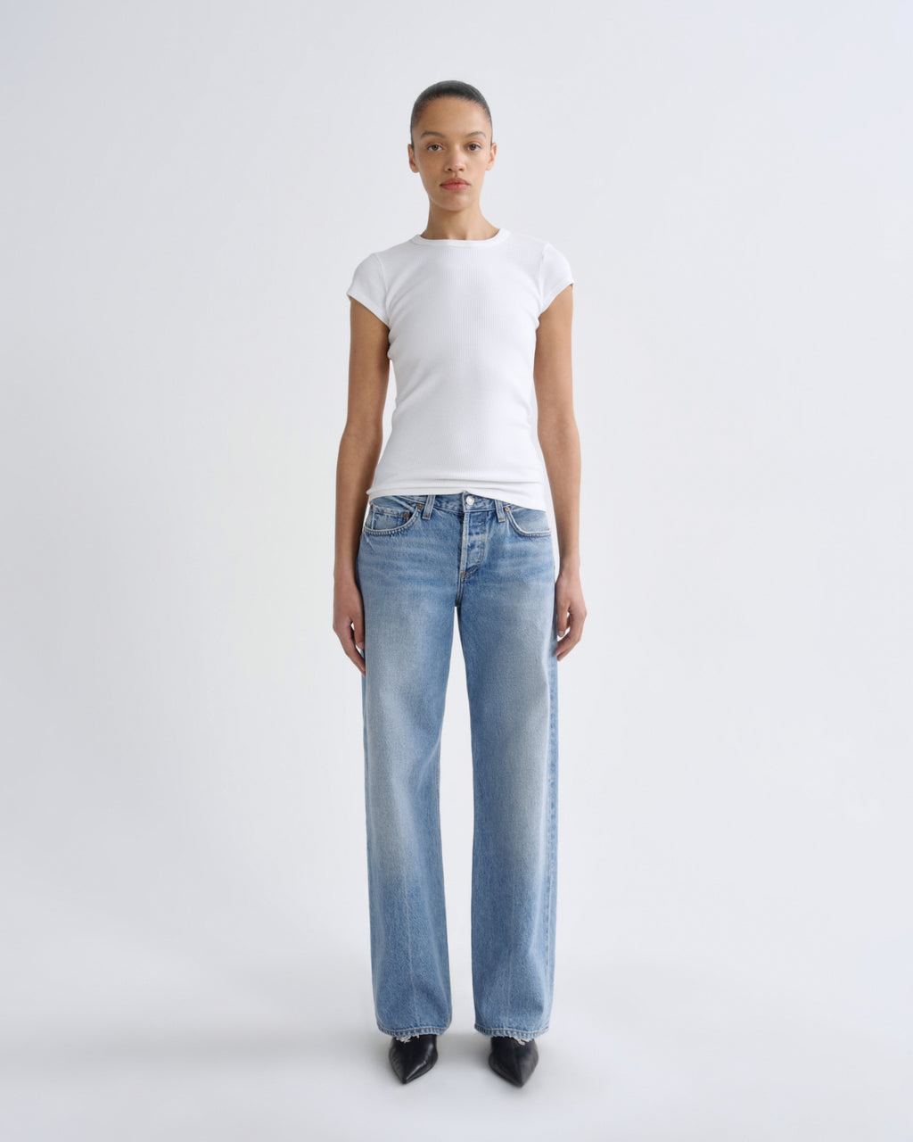 A model stands against a light gray studio background wearing a white short-sleeve crewneck t-shirt and light-wash blue straight-leg jeans with black pointed shoes.