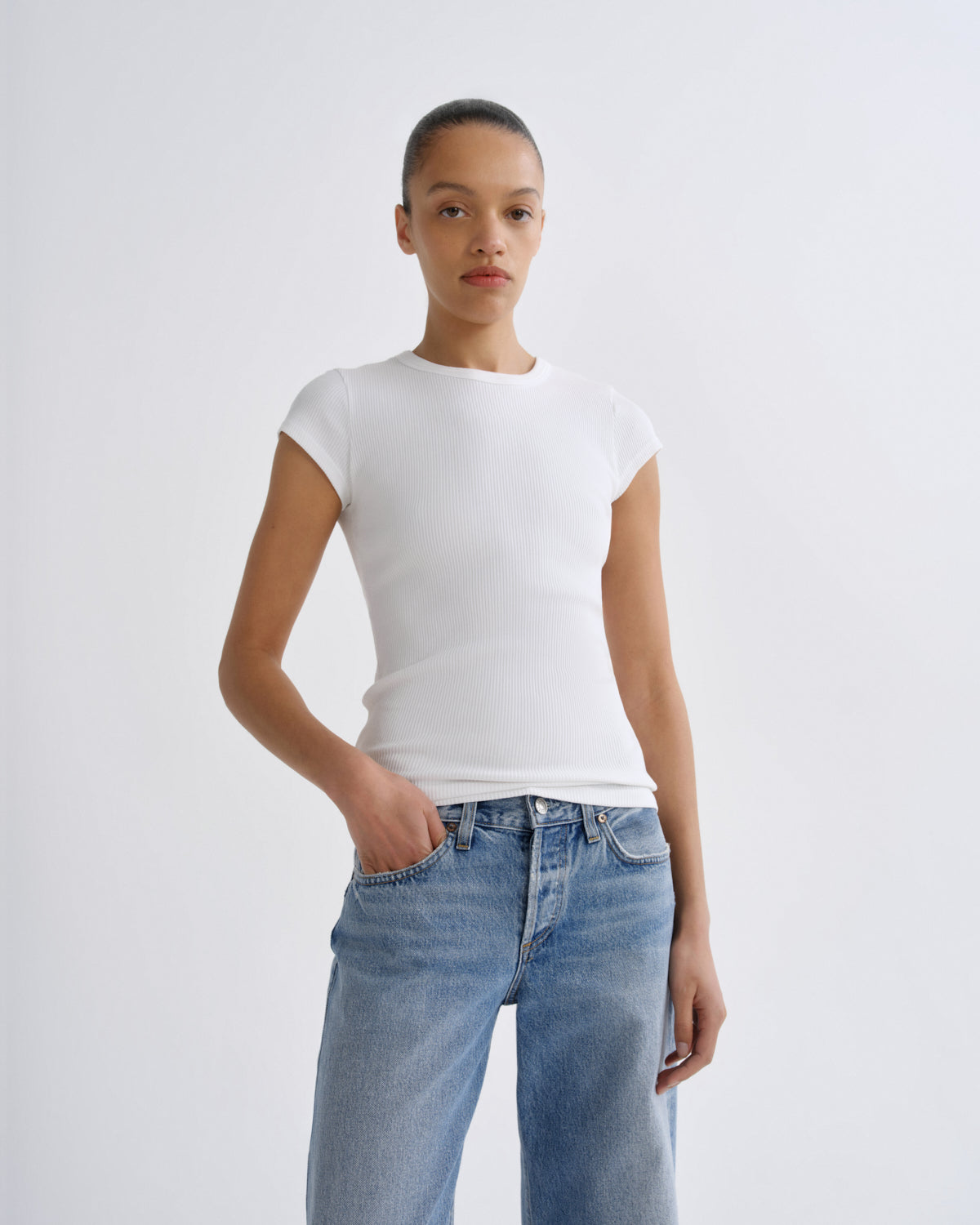 A model wearing a white short-sleeve crewneck t-shirt and light wash blue jeans, standing against a light gray studio background.