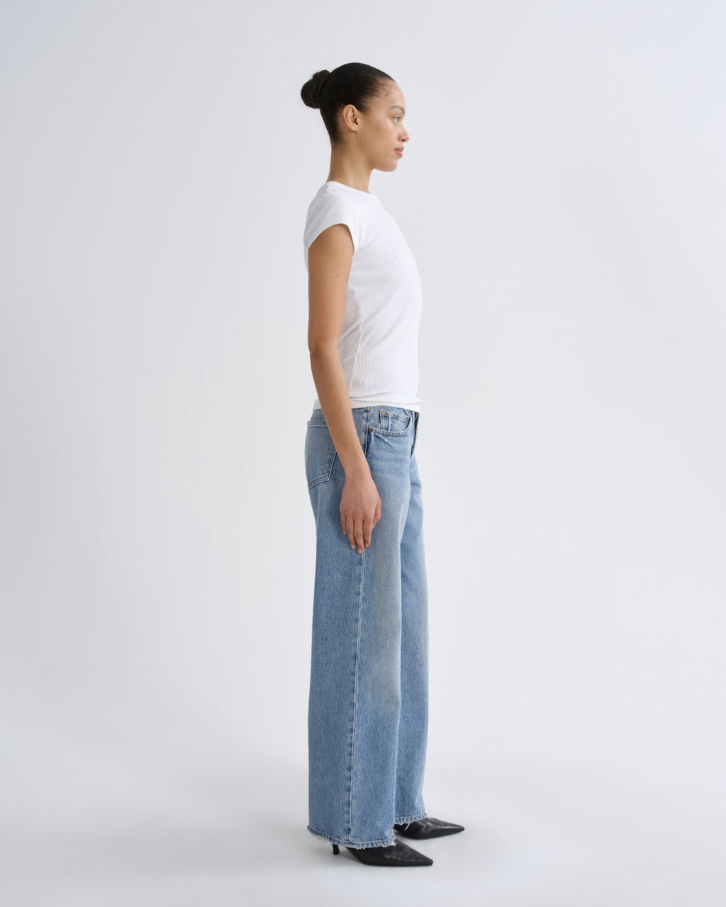 A woman in a white t-shirt and light-wash wide-leg jeans stands in profile against a plain light gray studio background.