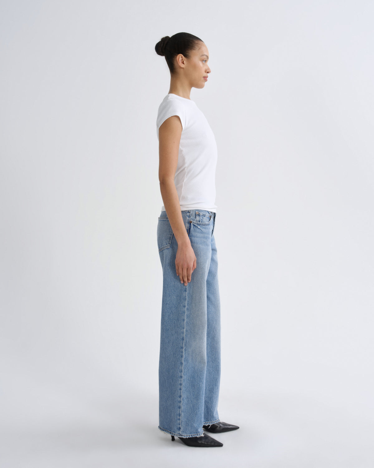 A woman in a white t-shirt and light-wash wide-leg jeans stands in profile against a plain light gray studio background.