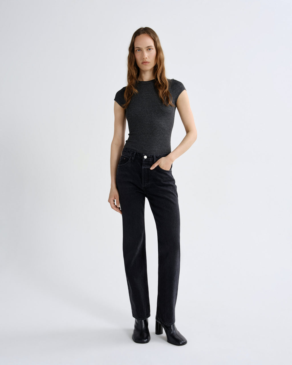 A woman modeling a dark gray short-sleeve knit top and black straight-leg jeans with black ankle boots, standing against a light gray studio background.