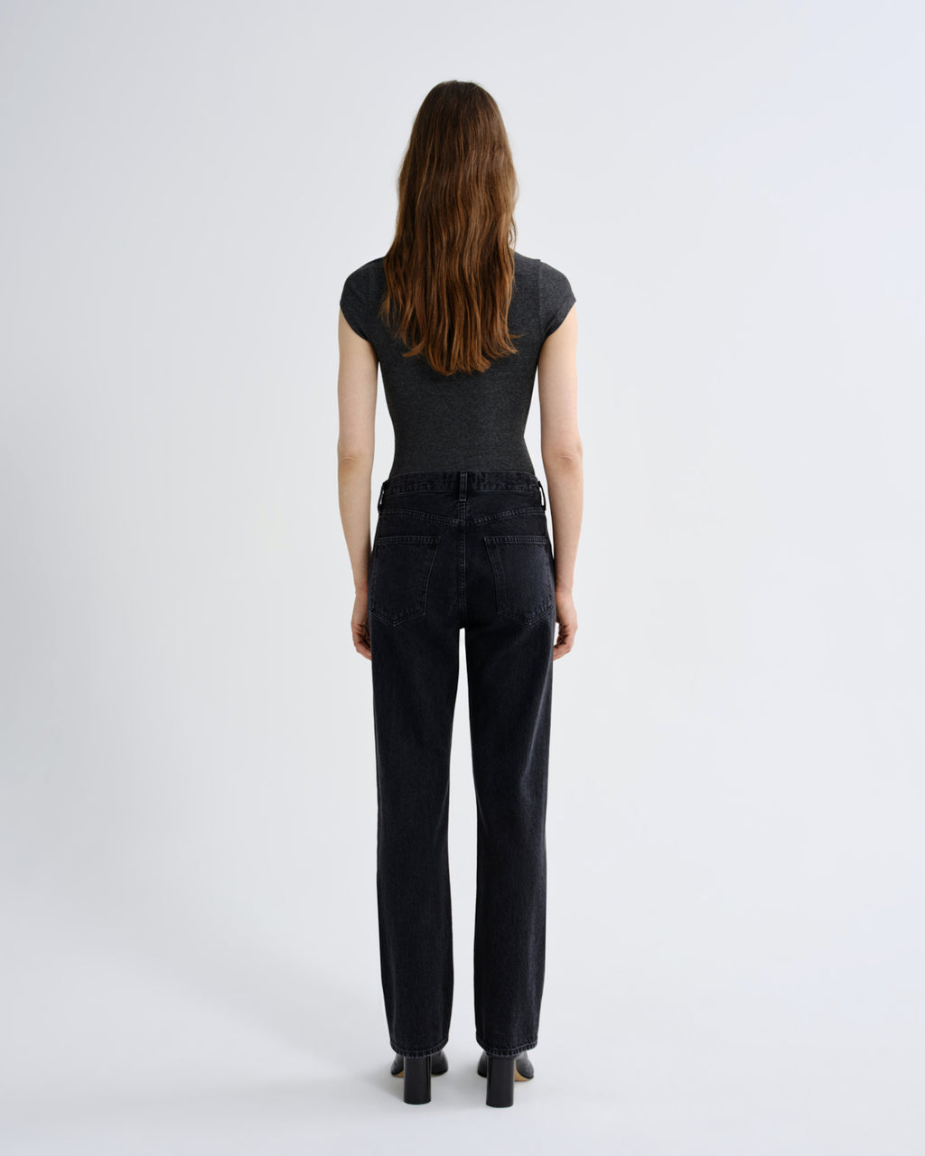 A woman viewed from behind wearing a dark gray short-sleeve top and black wide-leg jeans, standing against a plain light gray studio background.