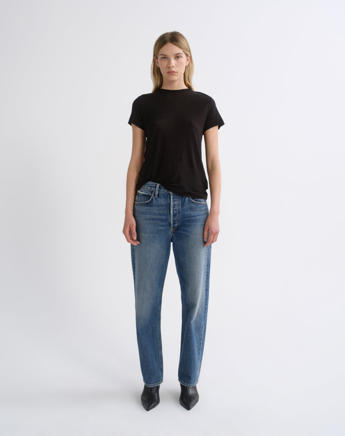 A woman stands against a light gray studio background wearing a plain black short-sleeve crew-neck T-shirt, blue straight-leg jeans, and black pointed shoes.