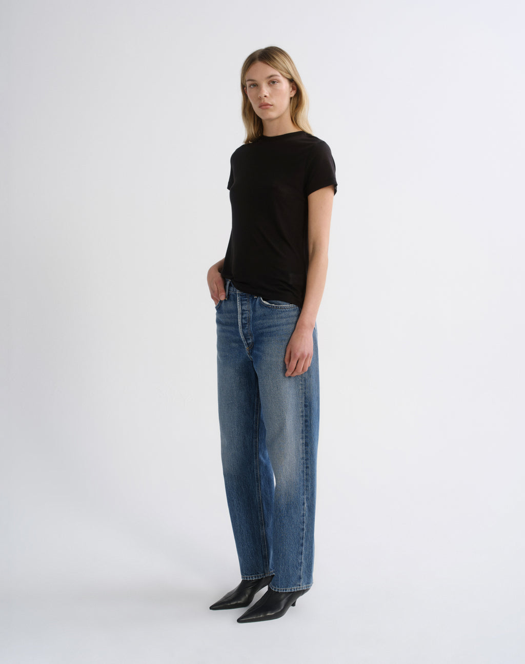 A woman modeling a black short-sleeve crewneck t-shirt and wide-leg blue jeans with black pointed-toe ankle boots, standing against a plain light gray studio background.