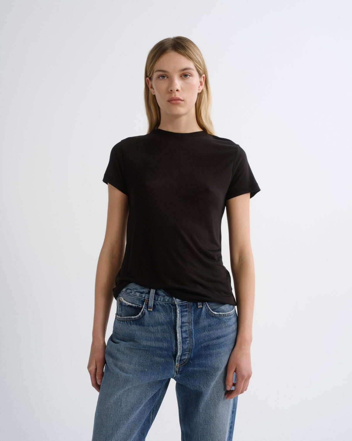 A woman modeling a plain black short-sleeve crewneck t-shirt paired with blue jeans, standing against a light gray studio background.