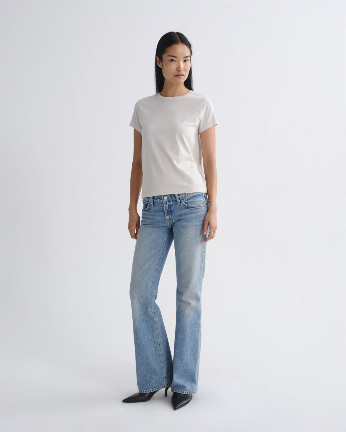 A model stands against a light gray studio background wearing a plain off-white short-sleeve crewneck t-shirt, light-wash blue flare jeans, and black pointed-toe heels.