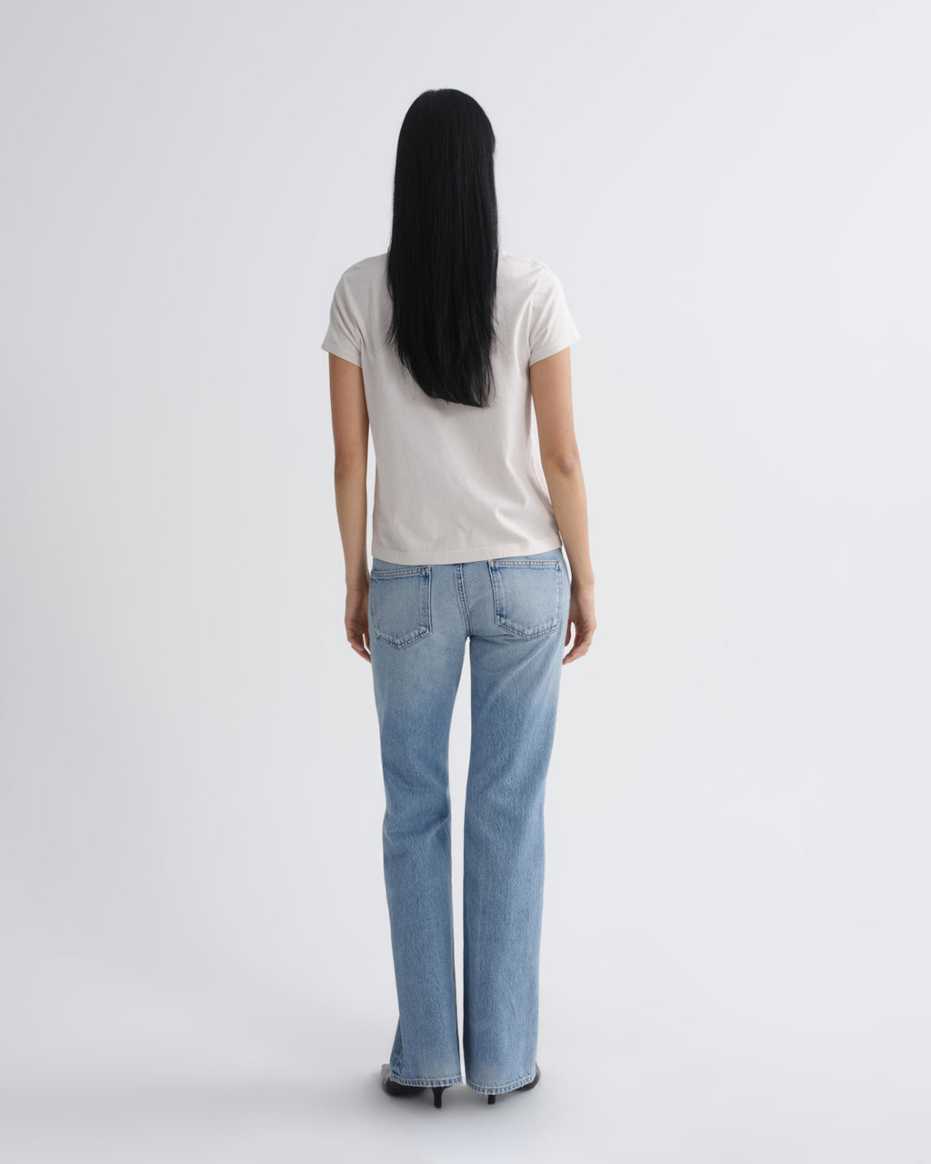 A woman with long black hair is shown from behind wearing a light beige short-sleeve t-shirt and light blue straight-leg jeans against a plain light gray studio background.