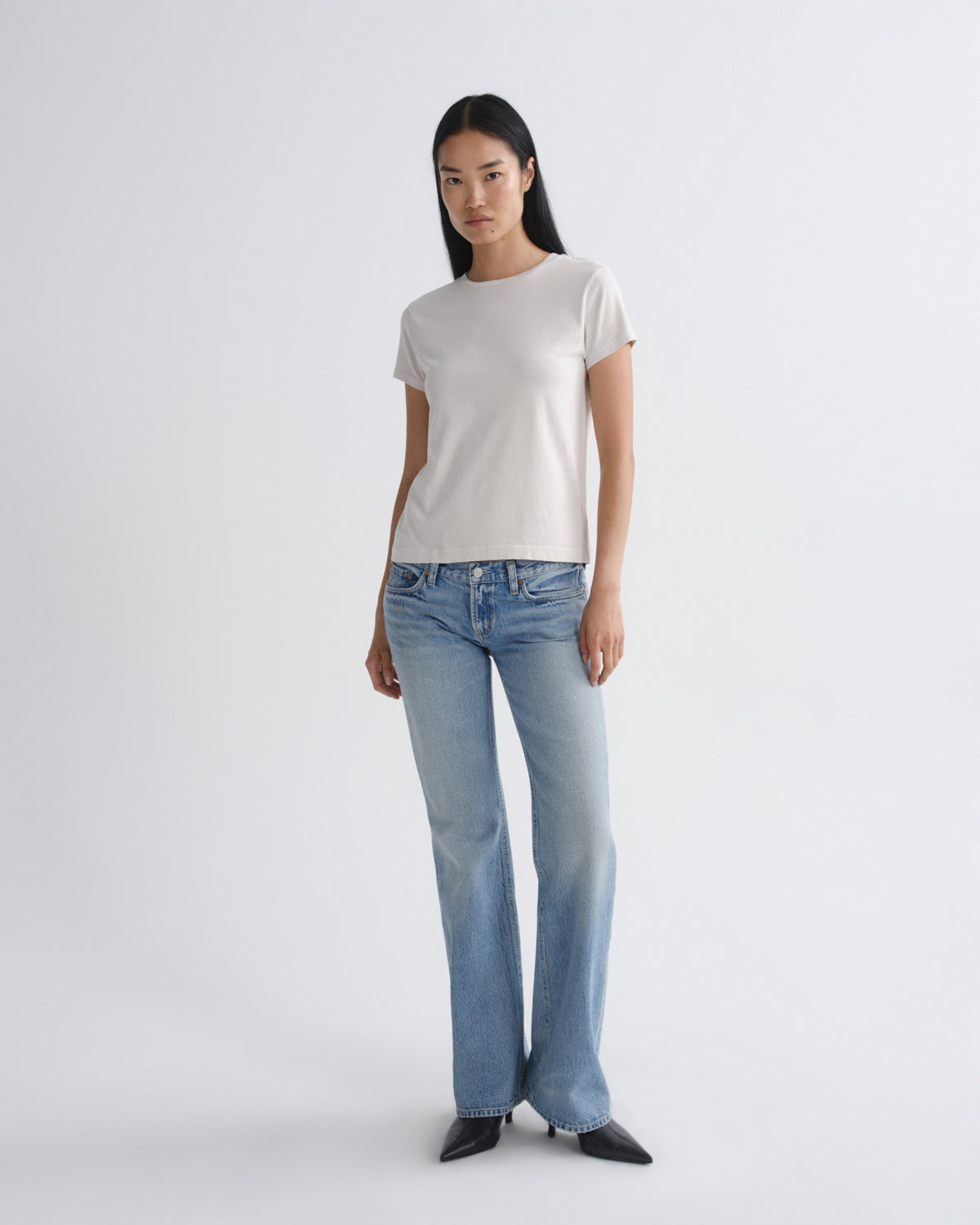 A woman modeling a light beige short-sleeve crewneck t-shirt and light-wash blue flare jeans against a plain light gray studio background.