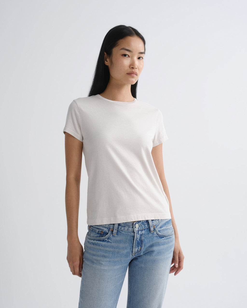 A woman modeling a plain white short-sleeve crewneck t-shirt paired with light blue jeans, standing against a light gray studio background.