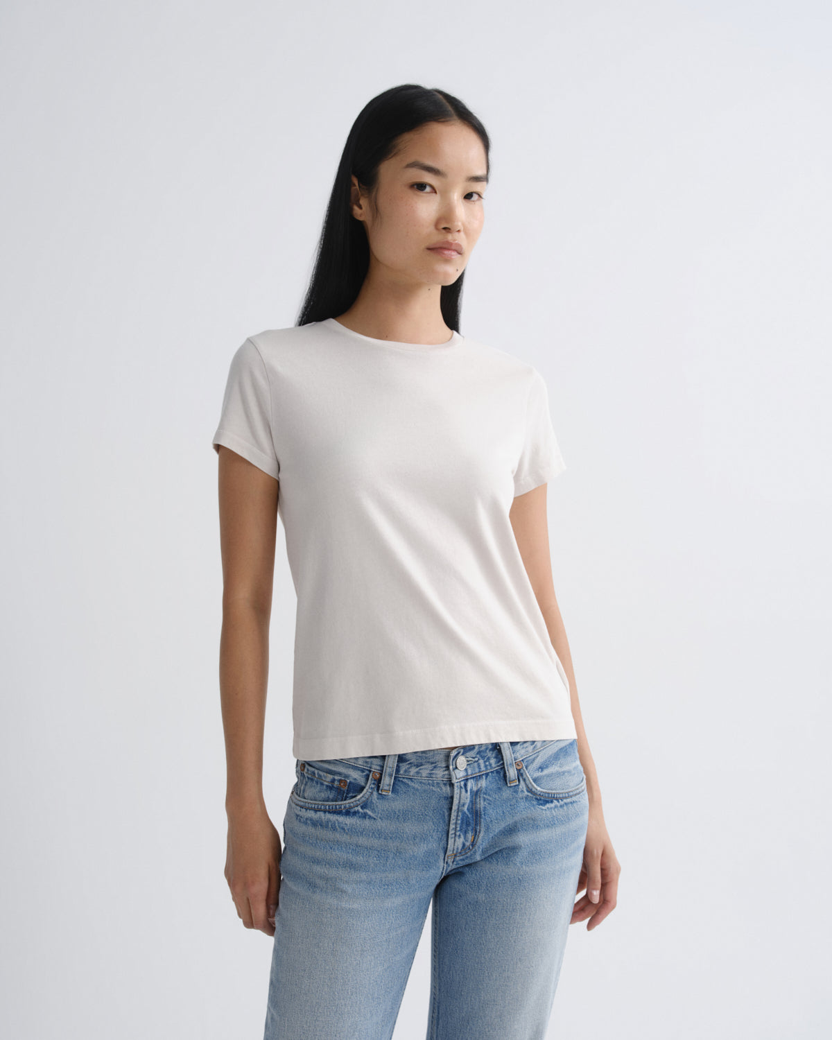 A woman modeling a plain white short-sleeve crewneck t-shirt paired with light blue jeans, standing against a light gray studio background.